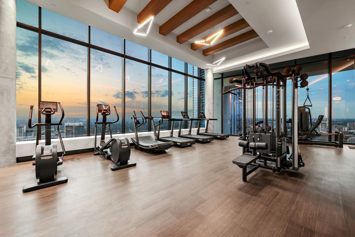 Modern gym with treadmills, exercise bikes, and weight machines, overlooking a cityscape at sunset through large windows.