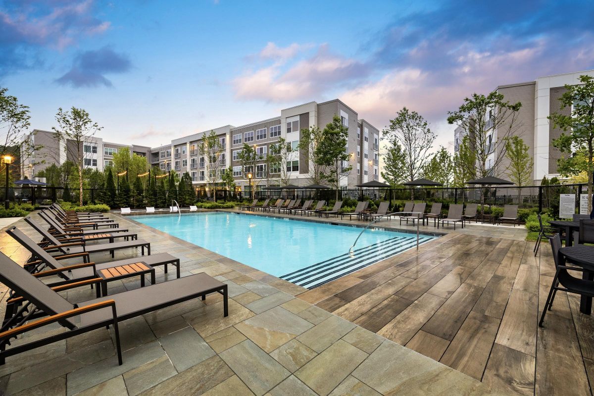 Outdoor pool with lounge chairs and surrounding trees, next to a modern apartment building under a cloudy sky.