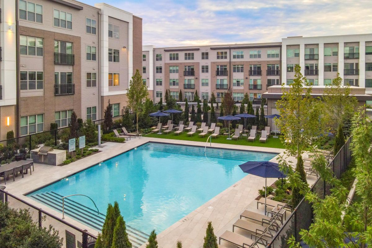 Apartment complex courtyard with swimming pool, surrounded by lounge chairs, umbrellas, and landscaped greenery.