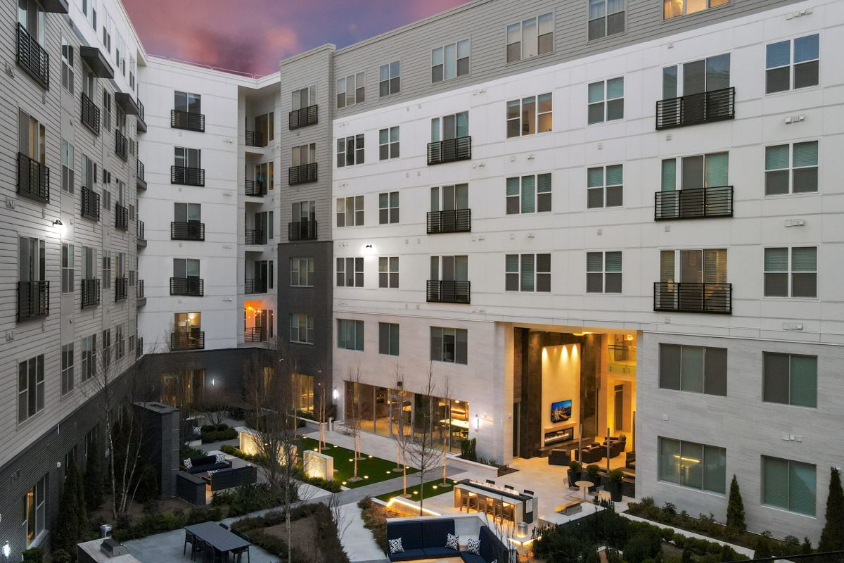 Modern apartment complex courtyard at dusk with outdoor seating, greenery, and large windows lit from within.