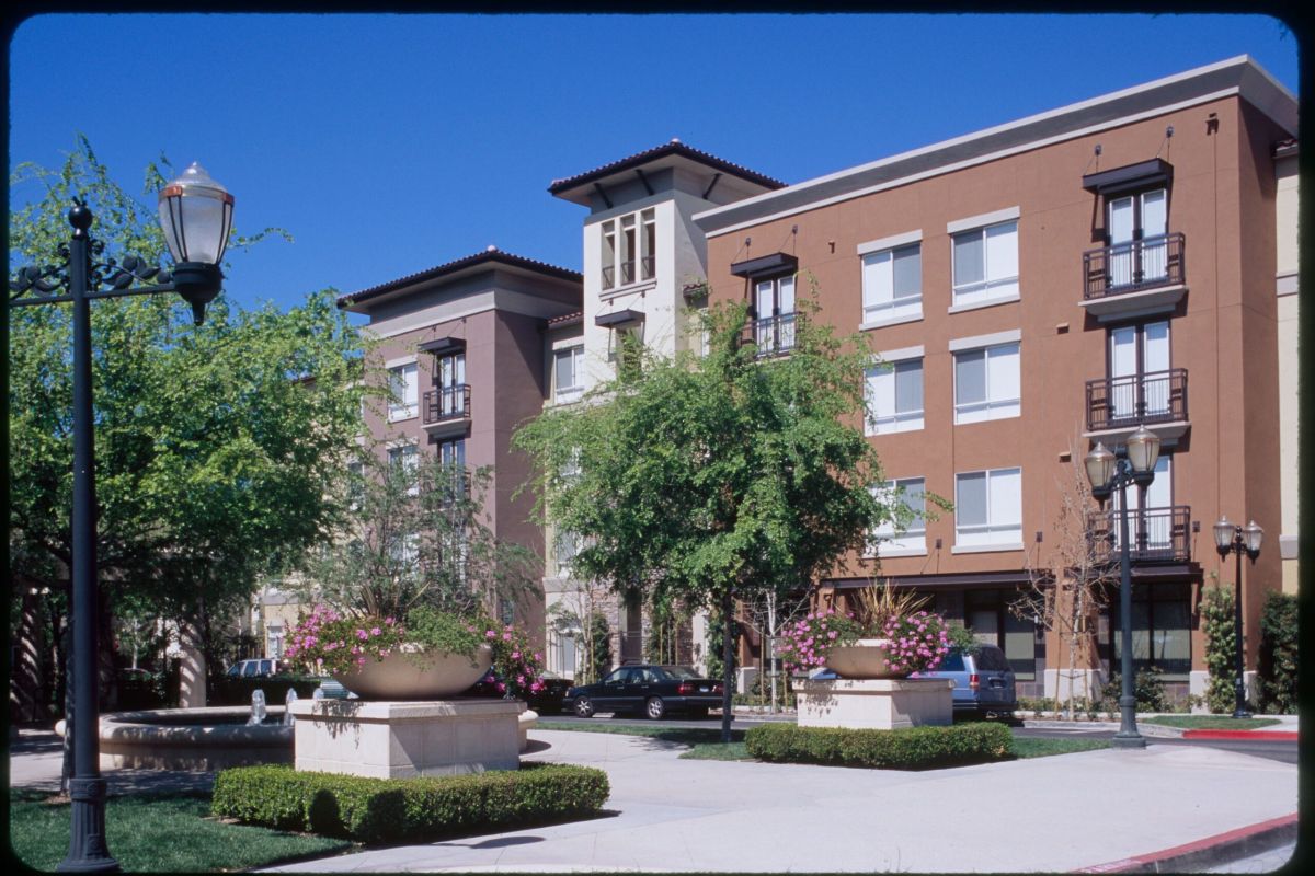Modern apartment building with balconies, trees, and flower planters under a clear blue sky.