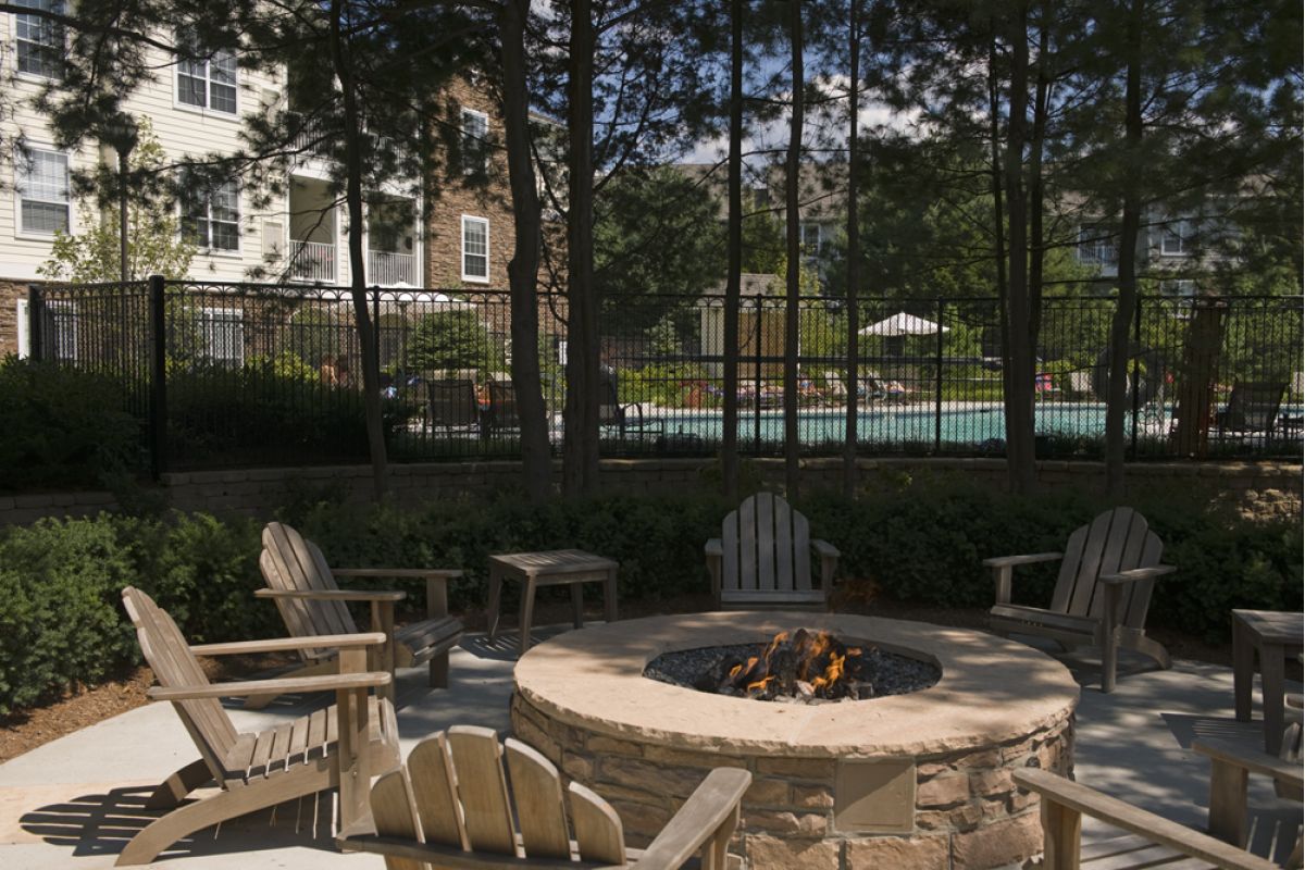 Wooden chairs surround a fire pit on a patio, with a pool and apartment buildings in the background.