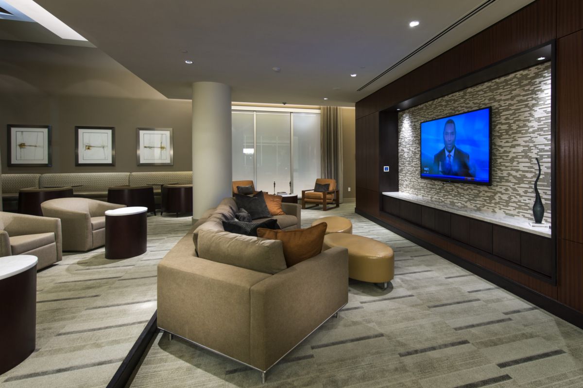 Modern lounge with beige sofas, armchairs, ottomans, and a wall-mounted TV showing a man in a suit.