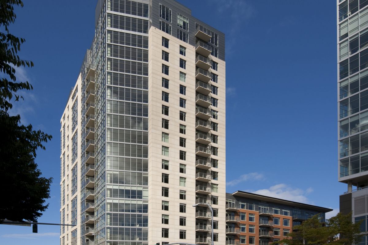 Tall modern apartment building with glass and stone facade, set against a clear blue sky on a city street corner.