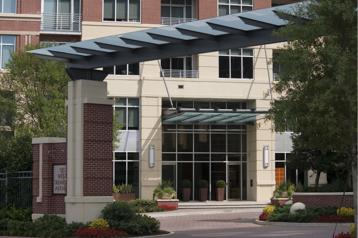 Modern apartment building entrance with glass canopy, brick columns, and landscaped greenery.
