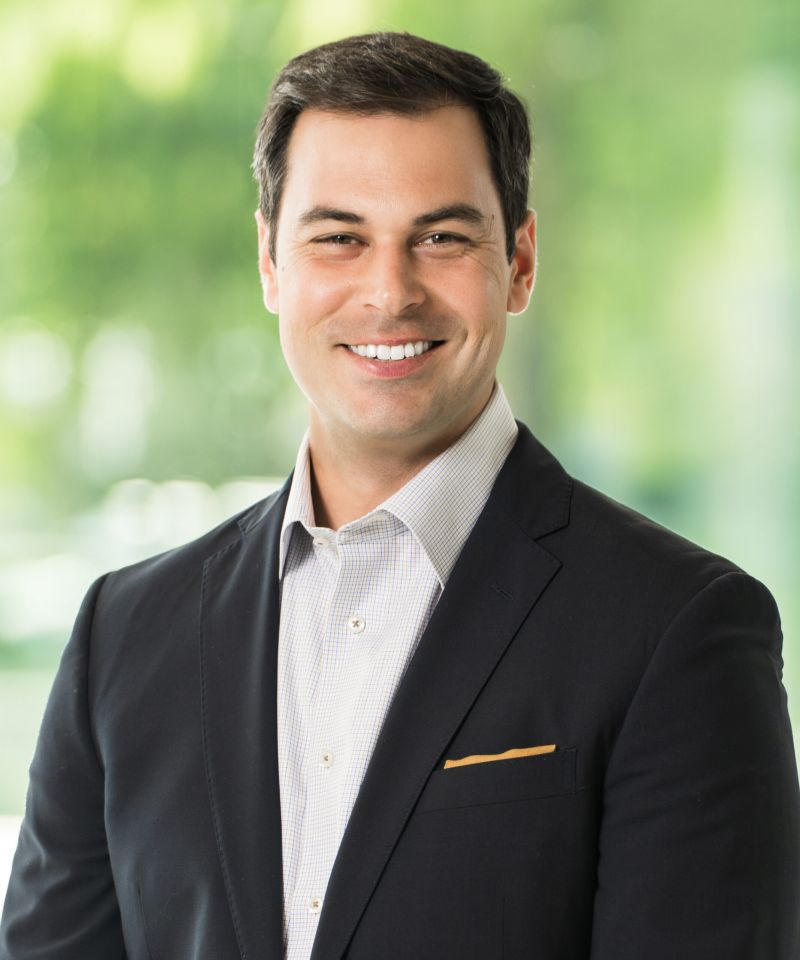 Smiling man in a dark suit and light shirt stands in front of a blurred green background.