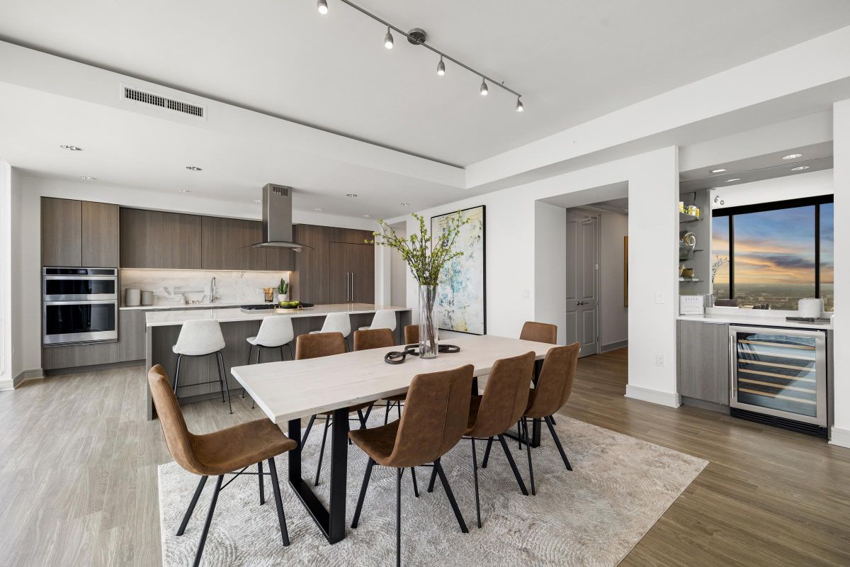 Modern open-concept kitchen and dining area with large table, brown chairs, and sunset view through floor-to-ceiling window.
