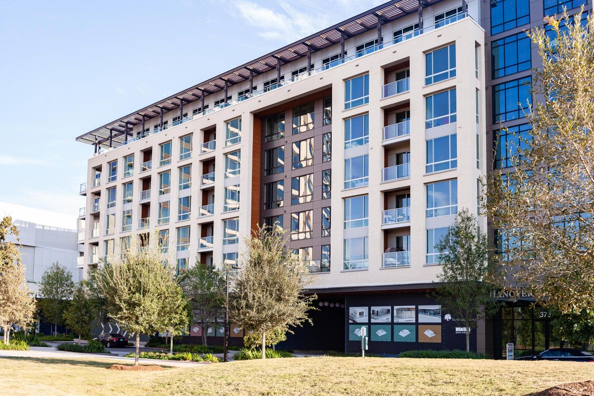 Modern mid-rise apartment building with balconies, large windows, and trees in the foreground.