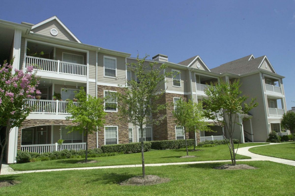 Three-story Churchill on the Park apartment building with balconies, green grass, trees, and pathways under a clear sky.