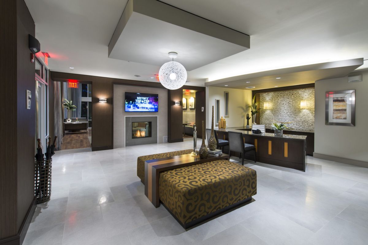 Modern hotel lobby with patterned seating, reception desk, TV, fireplace, and contemporary lighting.