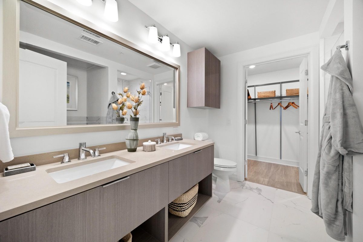 Modern bathroom with double sinks, large mirror, and view into a walk-in closet with shelves and hangers.