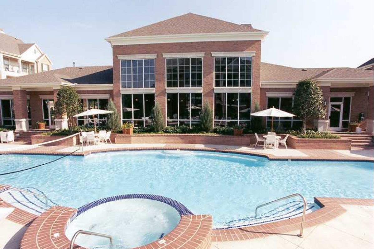Outdoor swimming pool and round hot tub at Churchill on the Park, set before a large brick building with tall windows and umbrellas.