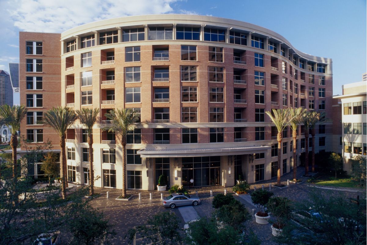 Modern multi-story brick building with palm trees and parked cars in front, seen under a blue sky.