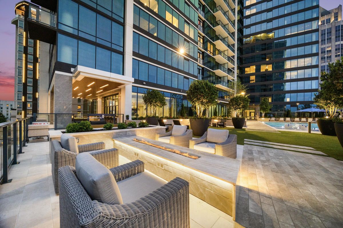 Modern outdoor lounge area with wicker chairs, fire pit, and pool, set beside a glass high-rise building at dusk.