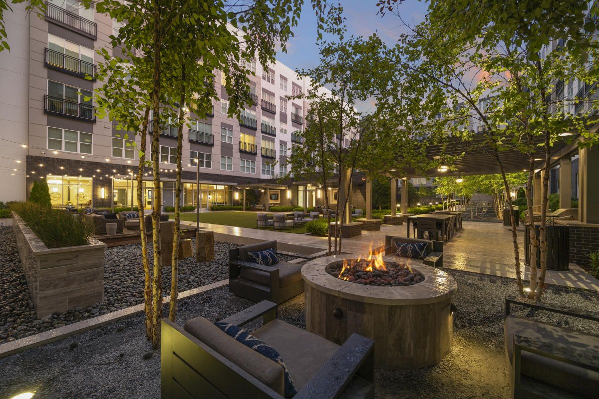Modern outdoor courtyard with a fire pit, seating area, trees, string lights, and apartment buildings in the background.