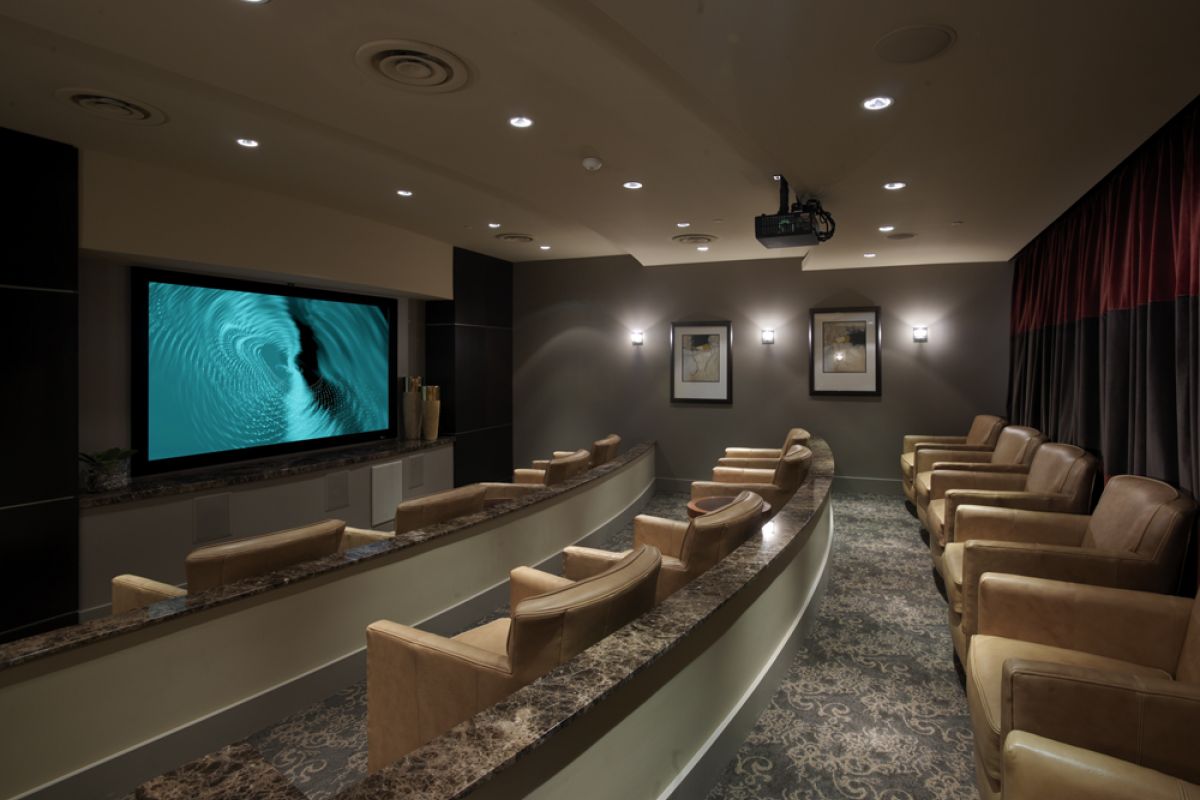 Small private theater room with rows of tan armchairs facing a large screen displaying a blue swirling image.