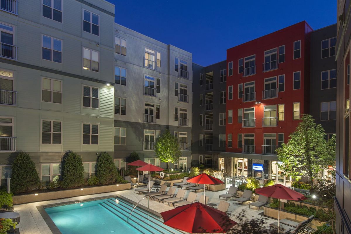 A modern apartment complex with a lit pool and red umbrellas at night.