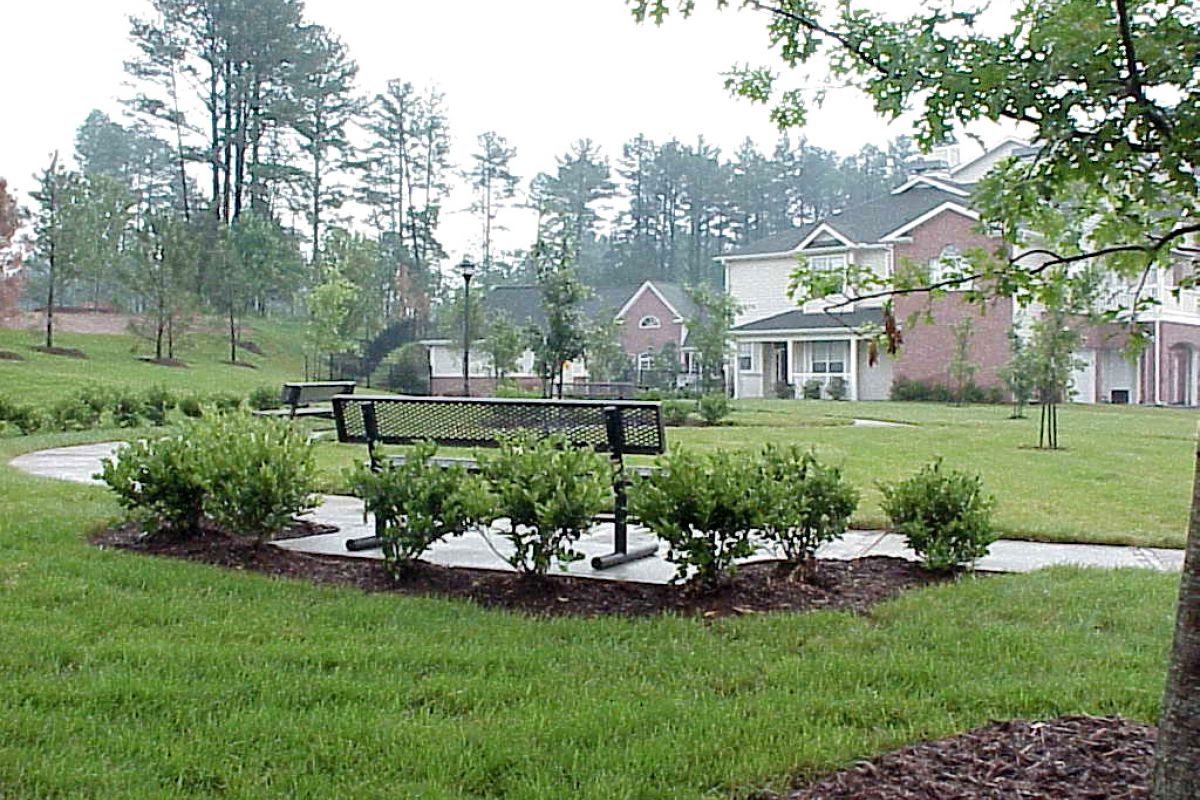 A park with a metal bench, shrubs, and pathways, surrounded by grass and houses in the background.