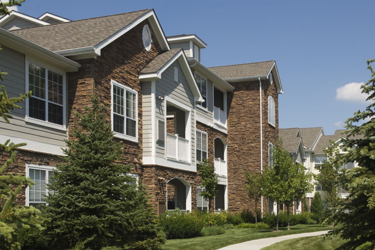 Modern apartment buildings with stone facades and balconies, bordered by green lawns and trees on a sunny day.