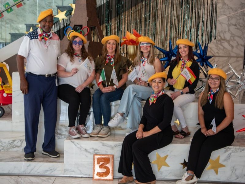 Seven people in yellow berets and scarves pose on decorated marble steps with Olympic-themed props.