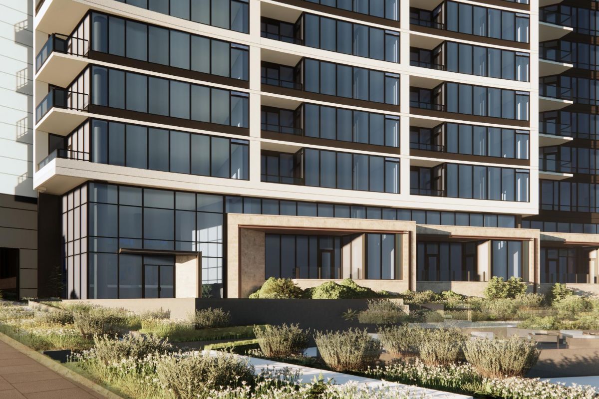 Modern glass building with balconies, surrounded by landscaped greenery and pathways, under a clear blue sky.