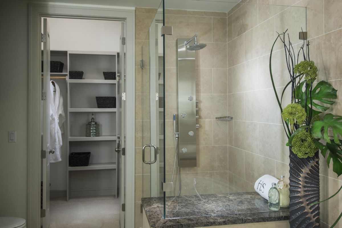 Hanover Company Modern bathroom with glass shower, large bathtub, and plants; door open to a walk-in closet with shelves and a robe.