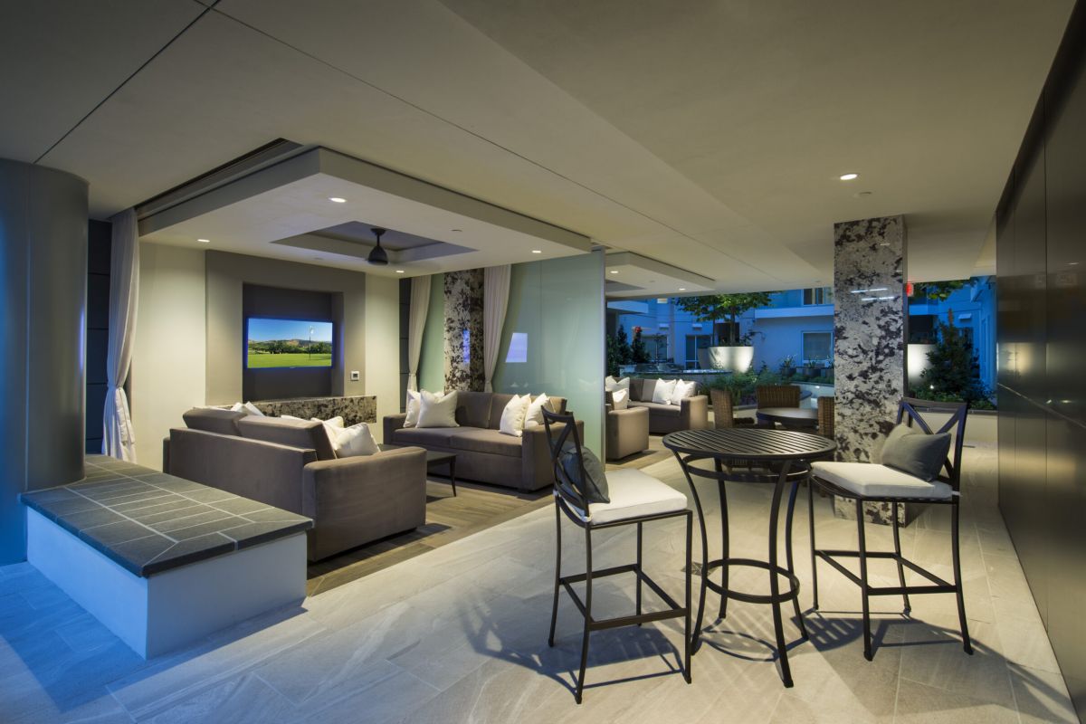 Modern outdoor lounge area with sofas, chairs, TV, and a tall table in a well-lit, covered patio.