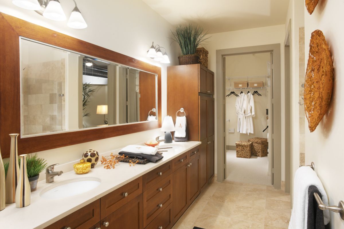 Modern bathroom with wooden cabinets, large mirror, bright lighting, and a walk-in closet with robes visible.