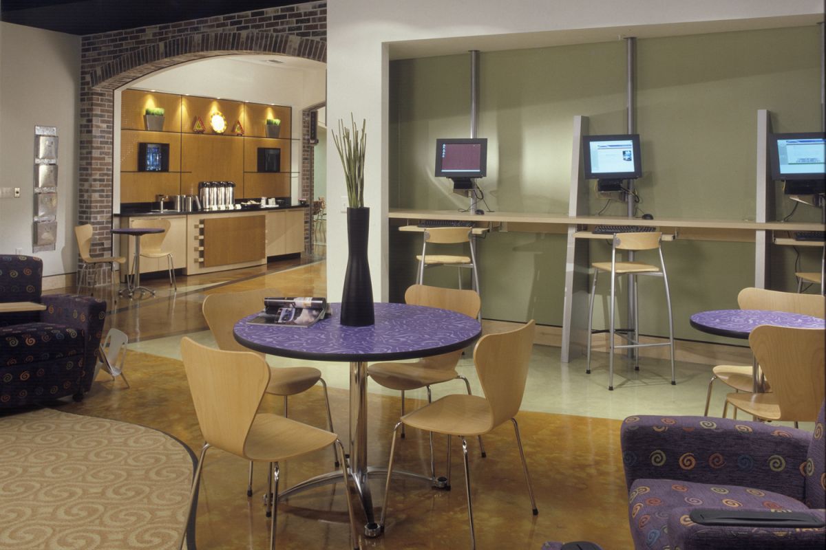 Modern lounge with tables, chairs, computer stations, and a refreshment area in the background.