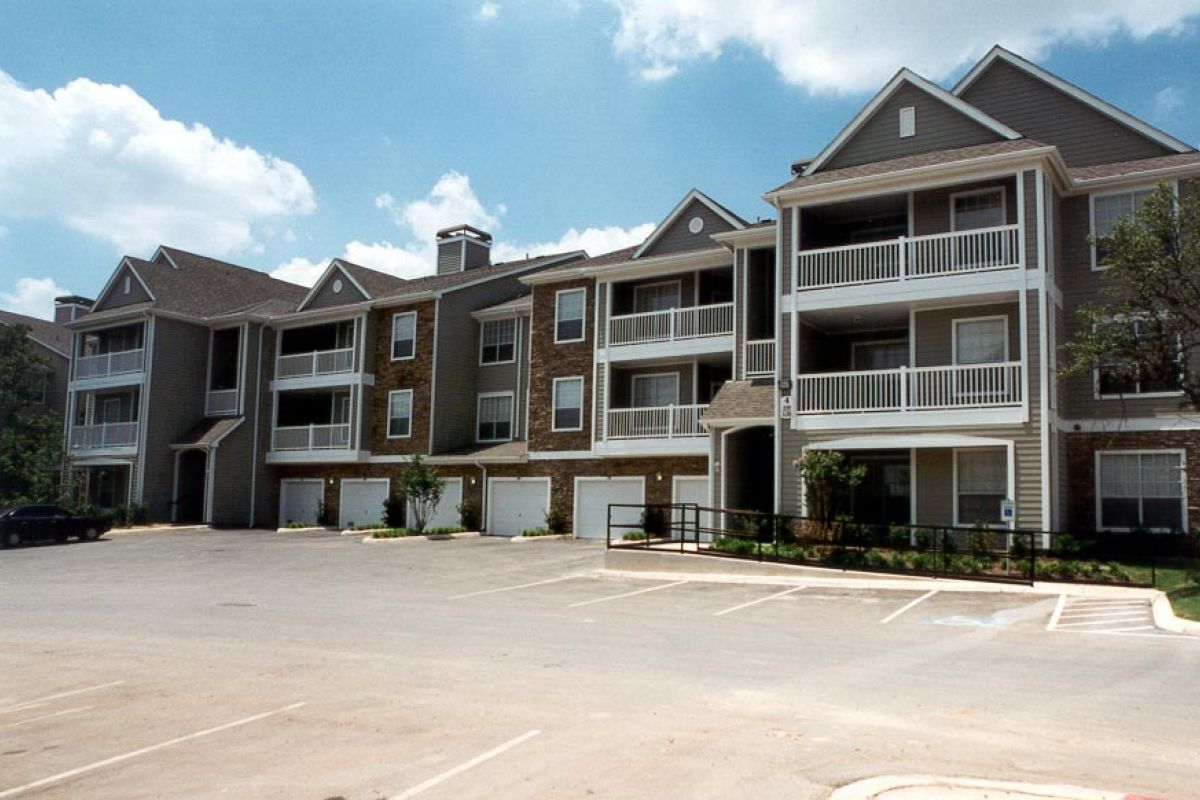 Three-story apartment building with balconies and garages, viewed from an empty parking lot on a sunny day.