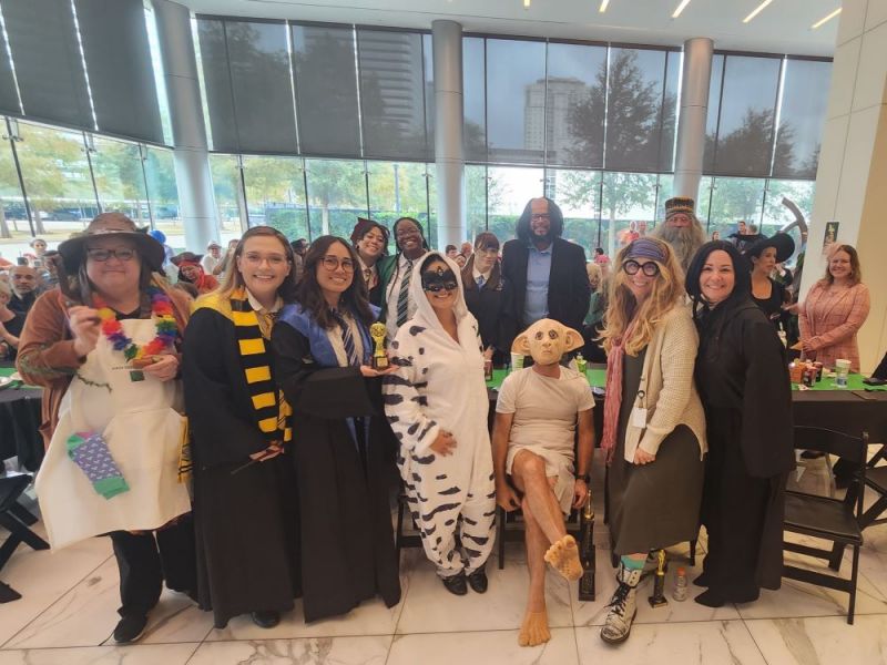 A group of people in costumes pose and smile together at an indoor event with tables in the background.
