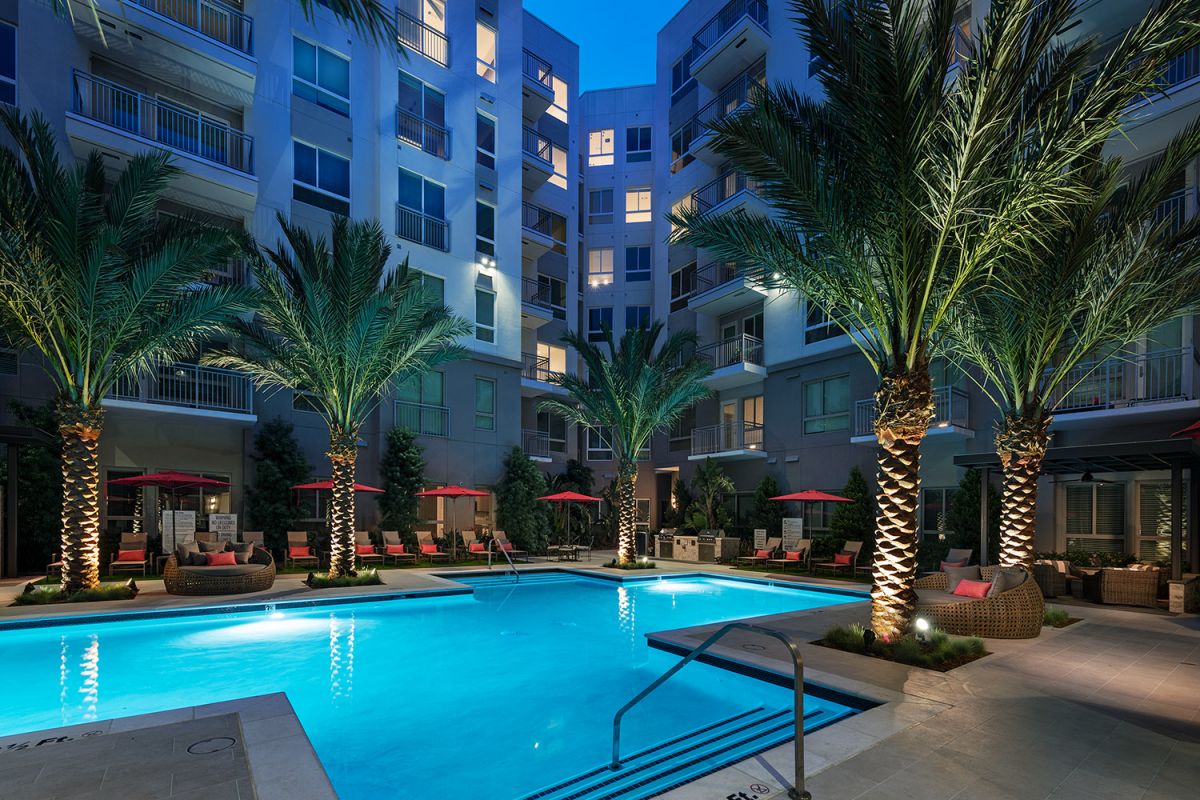 Modern Hanover Warner Center courtyard at dusk with a lit swimming pool, palm trees, and red lounge chairs.
