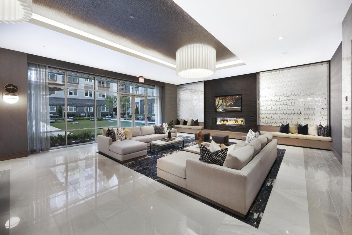 Modern lobby with beige sofas, a central coffee table, wall-mounted TV, and a fireplace. Large windows overlook a garden.