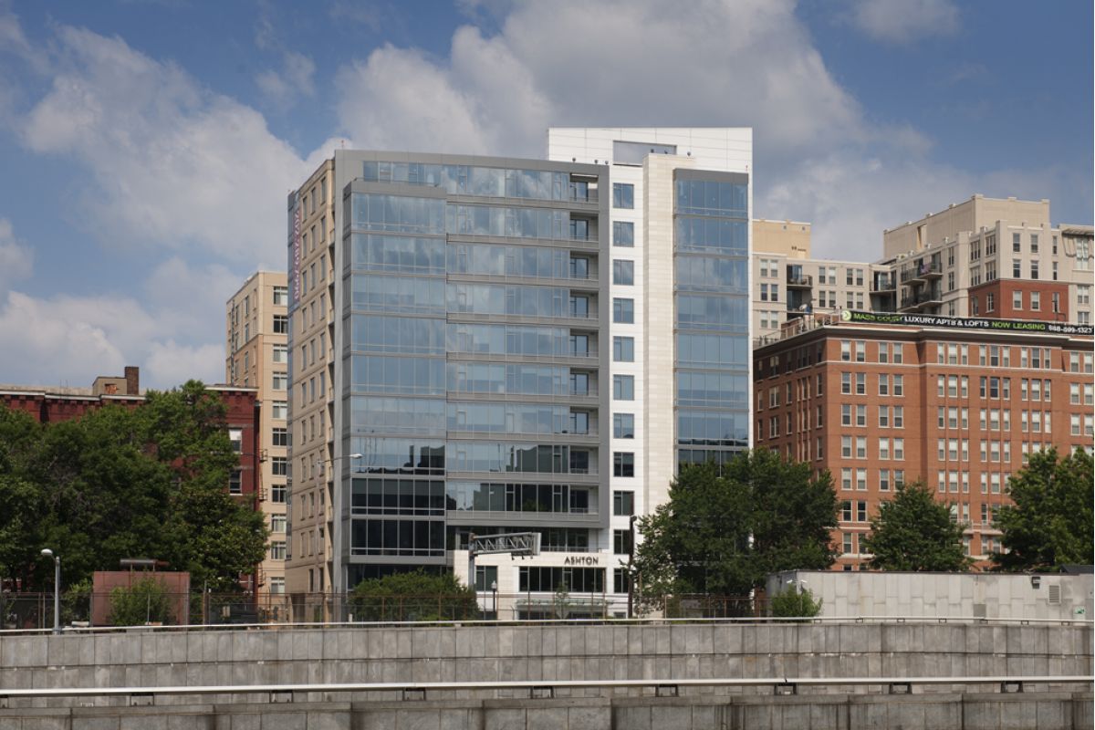 Modern glass office building with nearby brick buildings and trees under a partly cloudy sky.