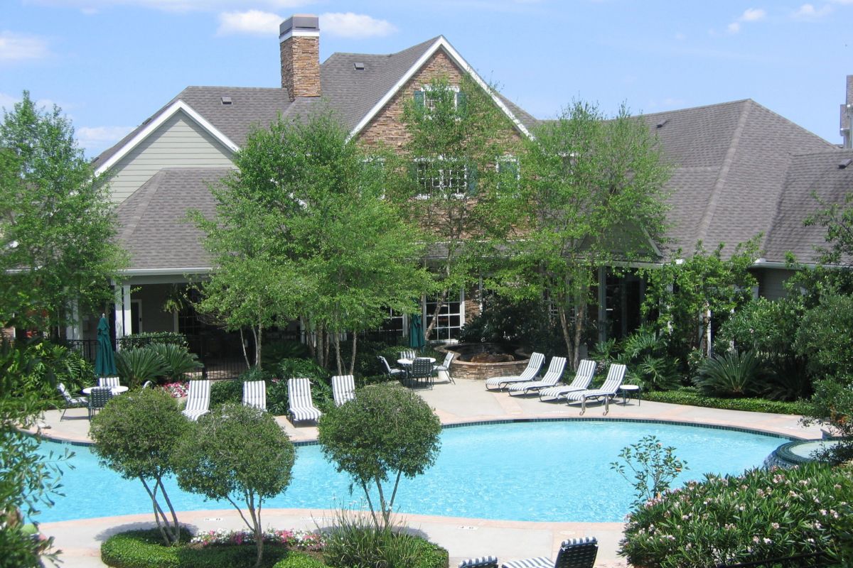 A backyard pool with lounge chairs and trees, reminiscent of the Broadwater Galveston, in front of a large house on a sunny day.