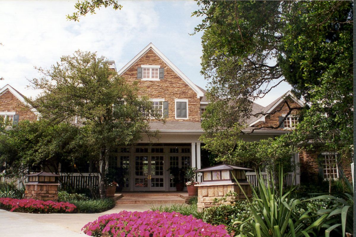 A brick house with a front porch, surrounded by trees, greenery, and vibrant pink flowers.