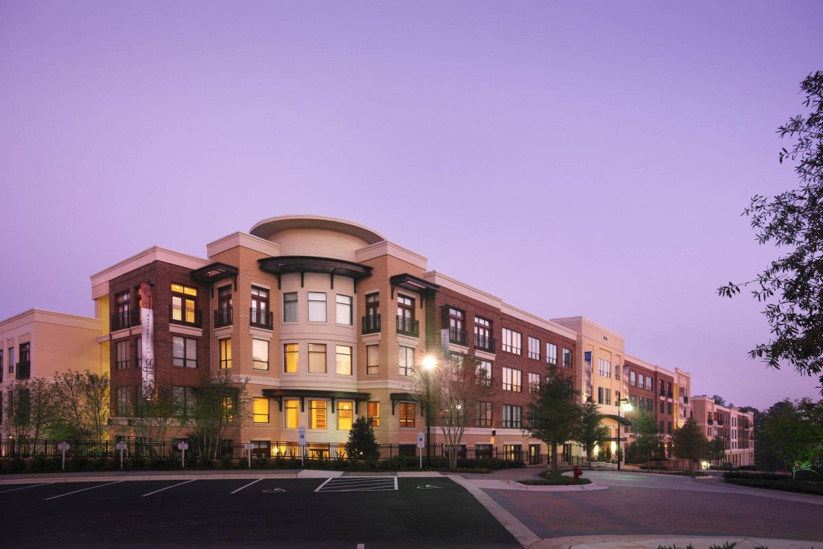 Modern apartment building at sunset with lights on and an empty parking lot in the foreground.