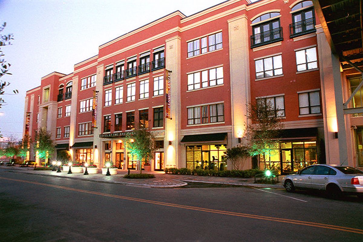 A modern, multi-story red brick building with shops and parked cars along the street at dusk.