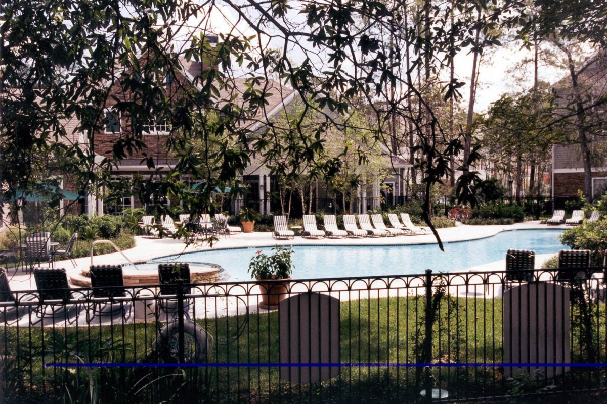 A fenced outdoor pool area with lounge chairs at Churchill on the Park apartments in Dallas, surrounded by trees and a building.