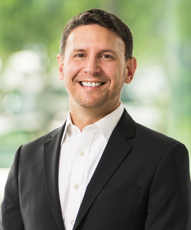 Smiling man in a black suit and white shirt, standing in front of a blurred green background.