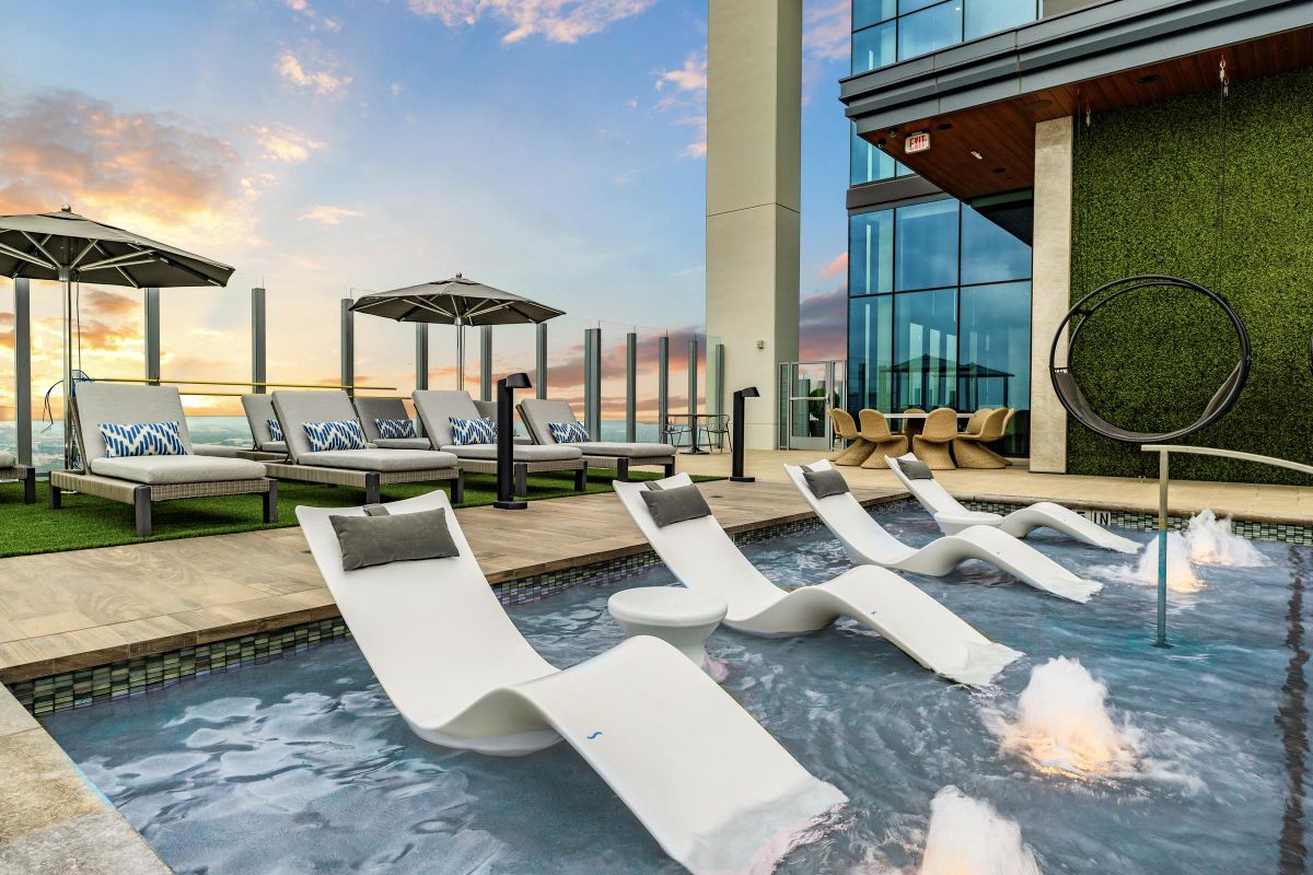 Rooftop pool with white lounge chairs, umbrellas, and sunset view. Modern building and seating area in the background.