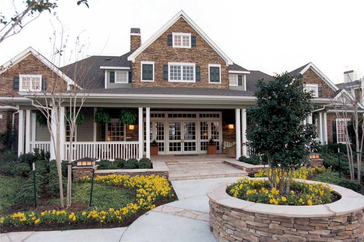 Large stone house with a wide porch, dormer windows, and flower beds with yellow flowers in front.