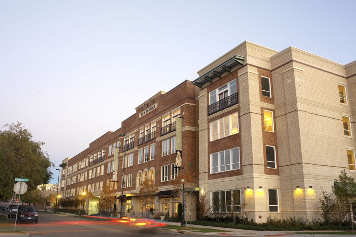 Four-story brick and beige building on a street corner at dusk with lights on and blurred car lights passing by.
