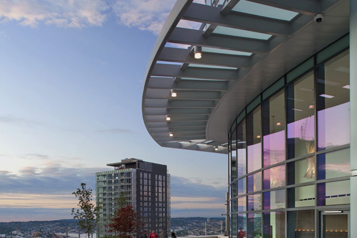 A modern rooftop patio with people sitting, city buildings, and a plane in the sky at sunset.