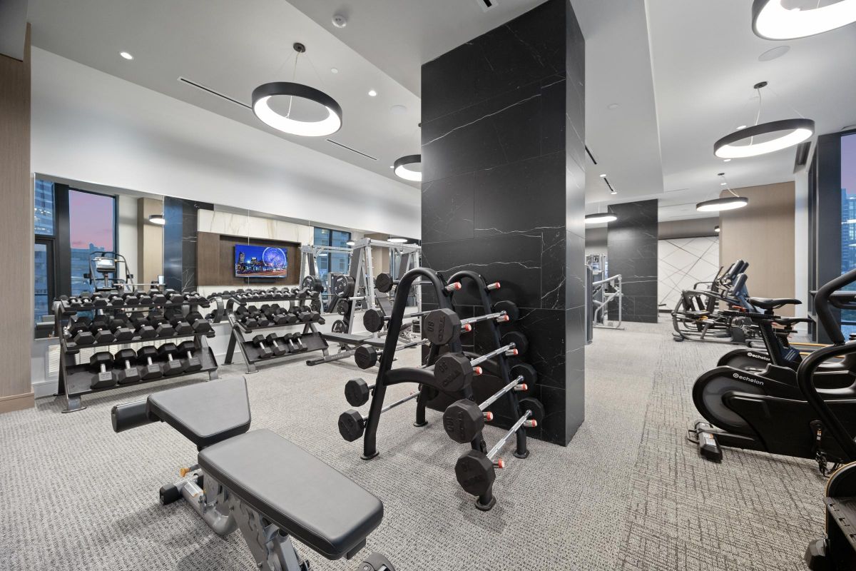 Modern gym with weights, benches, bikes, and TV on the wall. Floor-to-ceiling mirrors reflect exercise equipment.