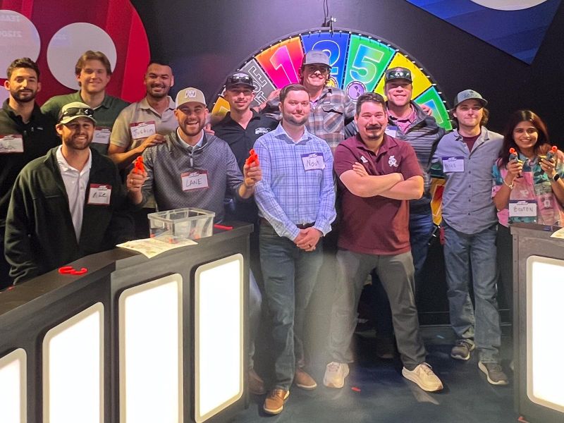A group of people pose and smile together in front of a colorful game show set with a spinning wheel.