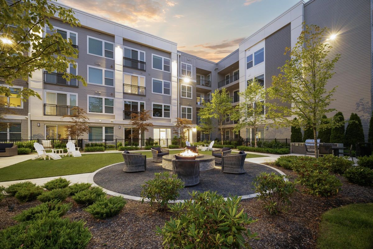 Apartment courtyard with fire pit, surrounded by chairs, trees, and greenery. Modern building in the background.