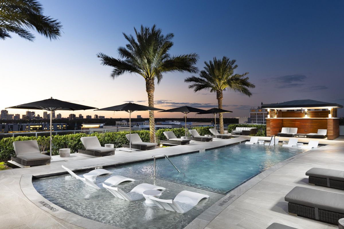 Rooftop pool area with lounge chairs, umbrellas, and palm trees at sunset, overlooking a city skyline.
