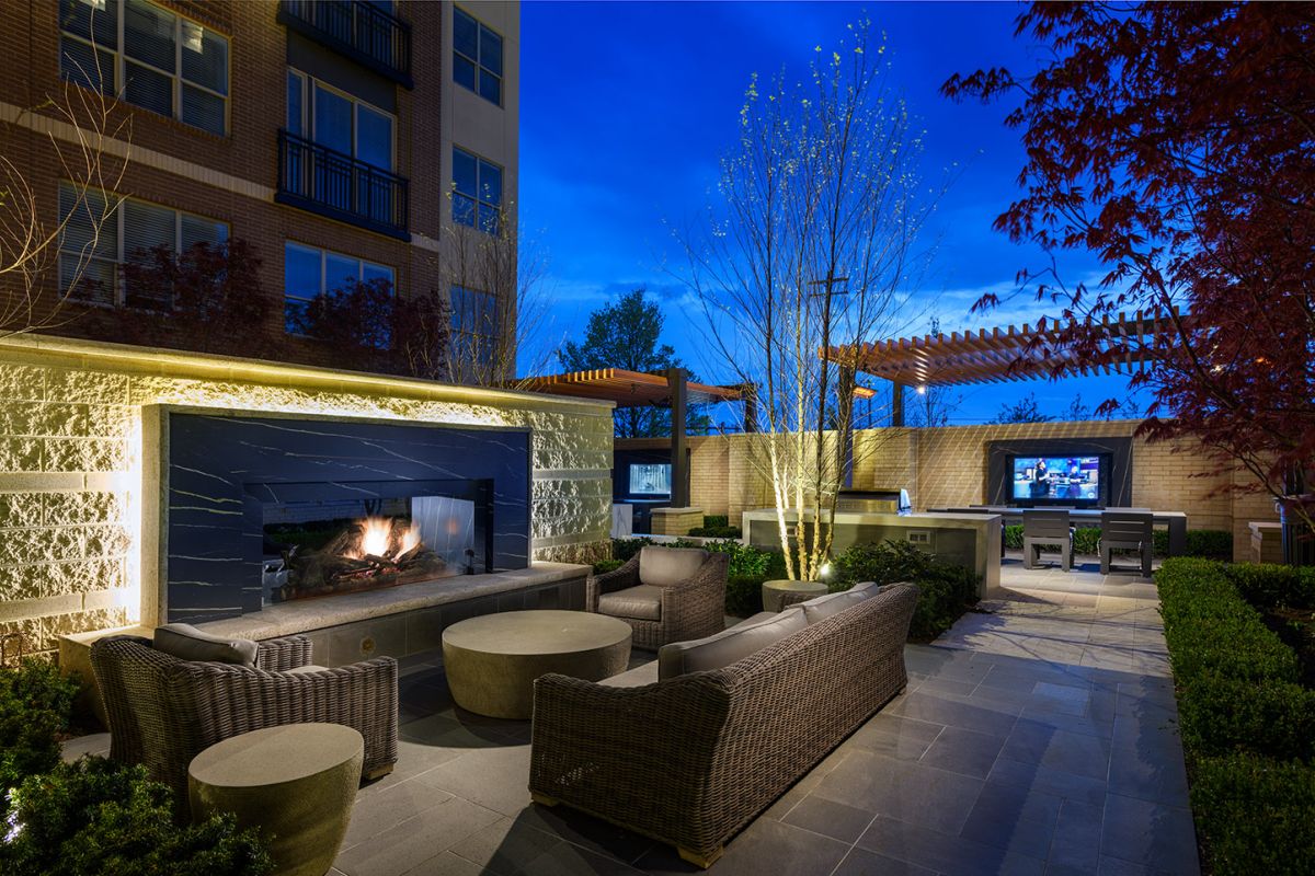 Outdoor patio at Hanover Exton Square features wicker furniture, a lit fireplace, and TVs under pergolas in a landscaped courtyard at dusk.