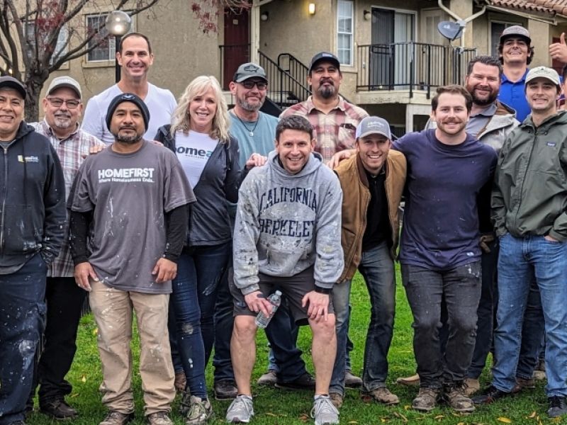 A group of sixteen adults smiling and posing together outside in front of an apartment building.